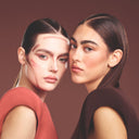 Due donne con capelli lisci ed eleganti posano vicine. Una ha linee di trucco di contorno sul viso e indossa un top corallo; l'altra indossa un top marrone con un trucco delicatamente sfumato. Sfondo marrone.
