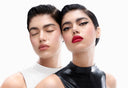 Due persone dai capelli scuri e lisci indossano top lucidi a collo alto, uno bianco e uno nero. La persona in nero ha un eyeliner alato ben marcato e il rossetto rosso; l’altra ha gli occhi chiusi e un trucco naturale.