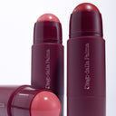 Three Too Much Attention - Stick Guance In Crema makeup sticks, featuring rounded pink tops and sleek burgundy cases, are arranged on a white background—two standing upright and one lying down—perfect for achieving un incarnato luminoso with a touch of blush cremosa.