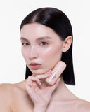 A woman with straight, dark hair and glowing skin holds a small bottle of Nudissimo Serum Fondotinta Satinato SPF30 close to her face against a plain white background. She has a neutral expression and minimal makeup.