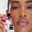 A woman with glowing skin holds two ALL IN ONE - BLUSH sticks—a pink and a bronze shade—close to her face, showcasing a natural, dewy viso look with shimmery lips and softly defined eyes.