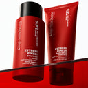 Un flacone rosso di Shampoo Riparatore e un tubetto abbinato di maschera per capelli appoggiati su una mensola rossa trasparente.