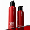 Due alte bottiglie rosse di Balsamo Ultra Riparatore Senza Risciacquo, dotate di erogatori neri, poggiano su una superficie rossa riflettente, mettendo in risalto le proprietà ristrutturanti per i capelli e la formula BOND ARCHITECT CPX per una riparazione profonda dei capelli fragili e danneggiati.