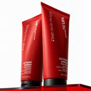 Due tubetti rossi di Maschera Riparatrice per capelli, con tappi neri, appoggiati su una superficie rossa traslucida.