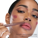 Una donna applica un FULLFACE SLIM STICK marrone - Matita multiuso in stick viso-occhi-labbra sulle labbra, con segni di trucco sulle guance e sulla fronte. Usa la tonalità 89 sulle guance e la tonalità 82 formula a lunga tenuta su viso e labbra.