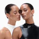 Two women with slicked-back hair and similar facial features pose closely. One wears a white high-neck top and has her eyes closed, as if enjoying the refreshing sensation of Crema Idratante. The other wears a black high-neck top and looks at the camera, her flawless skin suggesting the perfect base trucco achieved with this hydrating moisturizer.